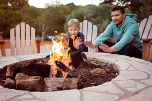 Tandem view of firepit and father and son, warming their hands by the fire in their yard