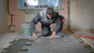 Repairman is renovating apartment wearing special clothes. He is laying a new tile on a levelled floor.